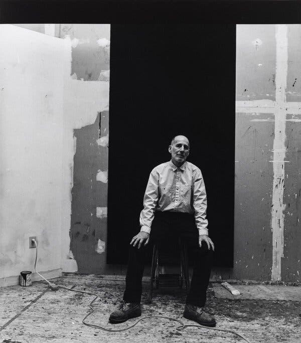 Robert Moskowitz, a balding slight man, in a black and white photo wearing a dress shirt and slacks while sitting on a stool, his hands resting on his knees, in front of a black panel pulled down over a wall covered in splotches of paint.