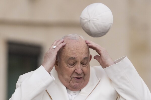 Pope Francis tries to catch his cap as wind blew it away while arriving for his weekly general audience in the St. Peter's Square at the Vatican, Wednesday, March 13, 2024. (AP Photo/Andrew Medichini)