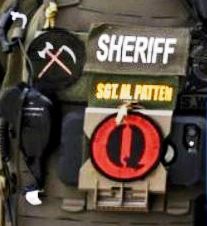 A zoom in on one soldier's uniform that has a patch with a black "Q" on a red background, and another that is a black field with an axe and scythe crossed over one another
