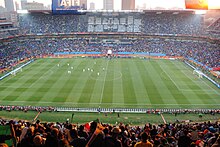 Un stade de football est pris en photo depuis le haut des tribunes. On aperçoit les spectateurs au premier plan, les joueurs au loin.