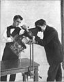 یک پزشک در حال نگریستن مستقیم به استخوان‌های دست یک بیمار با دستگاه پرتوافکن بدوی در سال ۱۹۱۰ میلادی.[پانویس ۴۹]