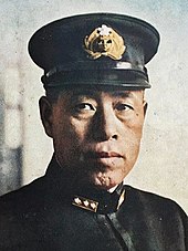 Černobílá kolorovaná fotografie bezvousého muže středního věku v černé uniformě. Na hlavě má černou brigadýrku se zlatým znakem.