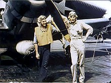 Barevná fotografie dvou mladých mužů pózujících před vrtulí letadla. Muž vpravo je oblečený do béžové letecké uniformy, na hlavě má leteckou kuklu.