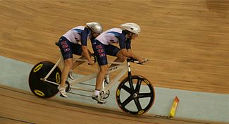 Dois atletas, usando capacete e uniforme dos Estados Unidos, pedalam na mesma bicicleta.