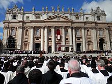 Bagian luar basilika pada hari yang cerah. Di latar depan, ratusan imam berjubah melihat ke arah podium di mana terdapat altar, dan sekelompok tokoh berjubah putih menghadiri Paus.