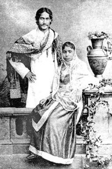 Black-and-white photograph of a finely dressed man and woman: the man, smiling, stands akimbo behind a settle with a shawl draped over his shoulders and in Bengali formal wear. The woman, seated on the settle, is in elaborate Indian dress and shawl; she leans against a carved supporting a vase and flowing leaves.