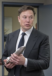 Musk, dressed in a suit, holds a metal model of the sleek Starship