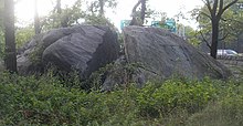 A photograph of a large rock, about the size of a small truck, that has a large fissure in the middle. The rock is surrounded by trees and other vegetation.