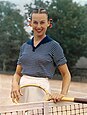 Eine Frau in gestreiftem T-Shirt und heller Hose steht auf einem Tennisplatz hinter dem Netz. Sie hält in einer Hand einen Tennisschläger und lächelt in die Kamera.