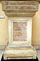 Das Foto zeigt einen Grabstein, auf seinem überkragenden Kopf und auf einem rechteckigen Textfeld sind Inschriften aus griechischen Buchstaben angebracht. Die Texte sind rot auf ockerfarbigem Stein.