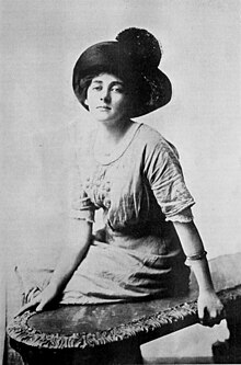 A young woman, seated on a bench, wearing a large dark hat, a light-colored blouse with a scooped neckline, and a light-colored skirt