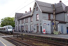 Foto eines grauen Bahnhofsgebäudes, im Vordergrund Schienen, auf denen sich ein Zug befindet