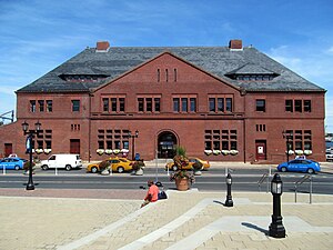 A three-story red brick railway station with a sloped roof viewed from an urban plaza