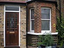 A brown brick building. Visible is a door at left, with a bowfront window at right; various tomato plants are also visible growing in front of the window.