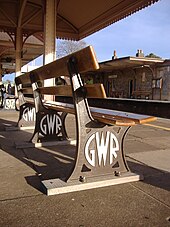 Un banco visto desde abajo y por detrás. Tres patas de hierro fundido pintadas de marrón tienen en bajorrelieve las letras "G W R" pintadas de blanco en un marco circular, y sostienen dos pares de tablones de madera que forman el asiento y el respaldo.