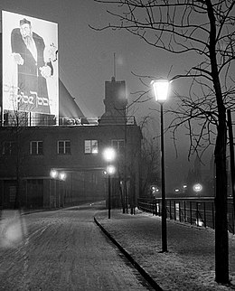Photographie en noir et blanc des quais de l'Isar à Munich. Au centre de l'image le bâtiment du Deutsches Museum est surmonté d'une large affiche éclairée présentant un stéréotype du Juif. Vue de la rive sur le bâtiment de la bibliothèque du Musée allemand, 1937.