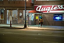 A woman in a brown fur coat stands smoking on the sidewalk of a city street, her hand resting on a pole supporting a red awning outside the entrance of a bar with a large red sign featuring the white words "Augie's"