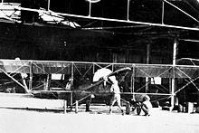 Front view of single-engined biplane and two men in front of a large open hangar