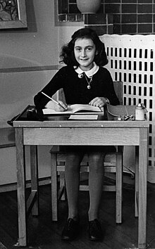 学校の机に座りノートを広げ、カメラ目線で微笑むアンネ・フランクの白黒写真。
