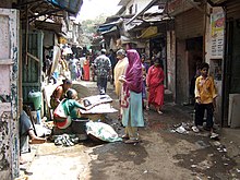 People walking on a narrow street. Shops and a stall are seen nearby.