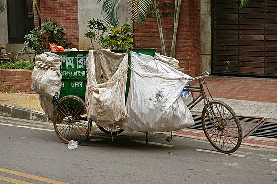 A transport rickshaw with a green container and additionally loaded with partially full plastic bags parked on the curb in front of a brick house