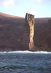 Visok pravokoten kup rjave skale stoji na sončni svetlobi pred obalo z visokimi pečinami, ki ležijo v senci.