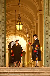 Frontale Farbfotografie von zwei Gardisten in einem Eingang zu einem Arkadengang mit einer Deckenlampe im Hintergrund. Der rechte Gardist steht seitlich mit einer Hellebarde. Beide tragen einen schwarzen Umhang über ihrer Uniform.