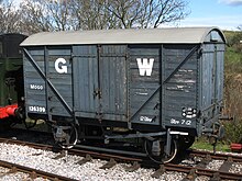 Un furgón de mercancías corto construido con tablones de madera. Los lados son de color gris medio con una gran G blanca a la izquierda de las puertas y una gran W blanca a la derecha.