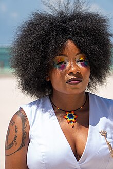 Photographie d'une personne noire portant un vêtement blanc, un collier en forme de fleur et du maquillage multicolore autour des yeux.