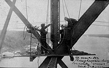 4 men working on the Poughkeepsie Bridge. The Hudson River is seen below with buildings along the shore making their height above the water apparent.