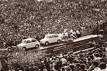 Foule assistant au tirage d'une loterie, le 2 septembre 1962 à Maribor (Yougoslavie). Exposés, les lots (visibles au centre de la photographie) sont des biens de consommation dont certains sont encore à l'époque peu accessibles dans le pays, par exemple : deux automobiles Zastava 600.
