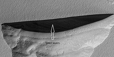Close view of wall of triangular depression, as seen by HiRISE layers are visible in the wall. These layers contain ice. The lower layers are tilted, while layers near the surface are more or less horizontal. Such an arrangement of layers is called an "angular unconformity".[269]