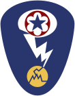 Oval shaped shoulder patch with a deep blue background. At the top is a red circle and blue star, the patch of the Army Service Forces. It is surrounded by a white oval, representing a mushroom cloud. Below it is a white lightening bolt cracking a yellow circle, representing an atom.