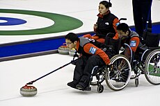 Homem em cadeira de rodas usa um suporte semelhante a uma vara para lançar a pedra. Um colega de equipe segura a sua cadeira e uma outra jogadora observa ao fundo.