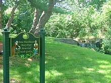 photograph of a multi-coloured carved wooden sign which reads "Anne Hutchinson/Mary Dyer Memorial Herb Garden, " behind which is a scenic small waterfall surrounded by green foliage.