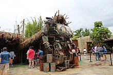 Une machine rouillée constituée de deux bras et de deux jambes devant une cabane en bois.