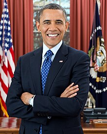 A portrait shot of Barack Obama, looking straight ahead. He has short black hair, and is wearing a dark navy blazer with a blue striped tie over a light blue collared shirt. In the background are two flags hanging from separate flagpoles: the American flag, and the flag of the Executive Office of the President.
