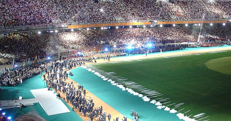 A portion of the stadium with stands full of people, a large artificial tree is on the right side of the image. A group of people are walking together on the stadium floor