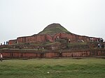 Ruins of a structure of red stone now resembling a small hill or mound.