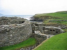 Polkrožna kamnita stena na levi namiguje na obstoj velike in starodavne zgradbe, na desni pa so ruševine različnih drugih kamnitih struktur. V ozadju nizka pečina ločuje vodno telo od travnatih polj.