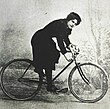 Seitenansicht einer jungen Frau mit dunkler Pluderhose und dunkler Bluse, auf einem zeitgenössischen Rennrad mit Herrenrahmen sitzend, das Gesicht der Kamera zugewandt