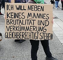 Bildfüllend ist ein Transparent aus braunem Karton mit der Aufschrift in Großbuchstaben: "Ich will neben keines Mannes Brutalität und Verkümmerung gleichberechtigt stehen".