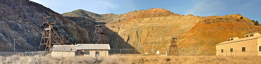 Panorama of fenced-off pit surrounded on three sides by rock walls. Three headframes and two buildings are within the enclosed area near the pit.