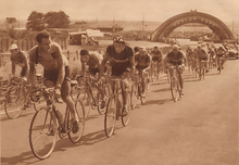Photo en noir et blanc d'un peloton de cycliste sur un circuit automobile. Deux coureurs au premiers plan s’observent.
