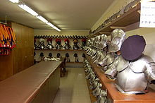 Farbfotografie eines Raumes mit Brustrüstungen und Helmen, die an beiden Wänden auf Regalen aufgereiht sind. Auf der linken Seite sind Schränke und Uniformen und in der Mitte steht ein langer Ausgabetresen.