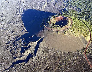 The conical volcano Lava Butte and its central crater are seen from above, with the volcano's right half forested while the opposite side is largely barren. An access road flanks the cone.