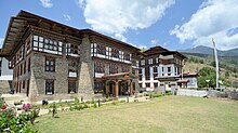 Ansicht des Gebäudes der Nationalbibliothek von Bhutan. Ein Gebäude mit einer hohen zweigeschossigen Wand aus gemauertem Naturstein und zwei Obergeschossen aus Fachwerk.
