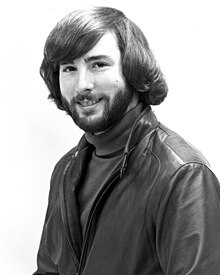 Portrait en noir et blanc, d'un homme barbu souriant, habillé d'un blouson en cuir.