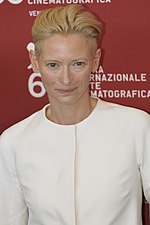 Upper torso of a female in her late forties standing in front of a red wall with white text and logos. She is wearing a white shirt