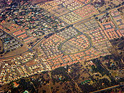 Bird's-eye view of residential neighborhoods. The houses at the bottom third of the picture have much larger yards and more trees. The neighborhood in the center of the picture has a very defined road shaped like a backwards 6, and the houses have orange and grey roofs and are very close to each other. The neighborhood at the top right has multicolor roofs. There is a road cutting through the middle of the picture.
